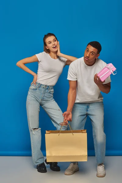 stock image positive multicultural couple buying present, holding shopping bag on blue backdrop, consumerism