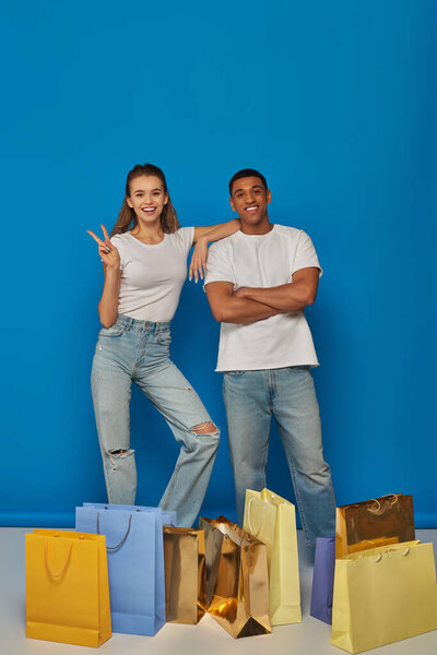 positive multicultural couple standing near shopping bags on blue backdrop, showing peace sign