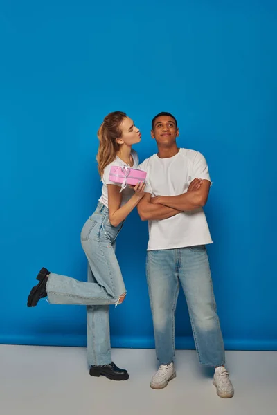 stock image beautiful woman holding present near happy african american man on blue backdrop, air kiss