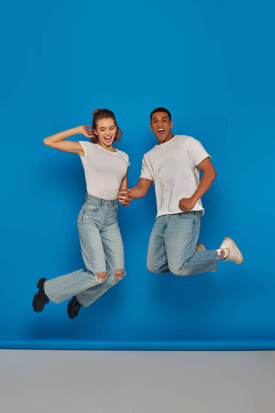 позитивная мультикультурная пара, держась за руки и прыгая на синем фоне, полный обзор длины