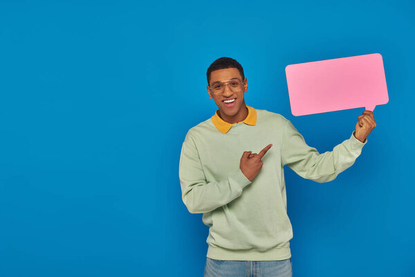 cheerful african american man in eyeglasses pointing at blank speech bubble on blue backdrop