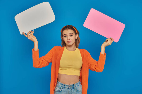 confused young woman in casual attire holding speech bubbles on blue background, space for text