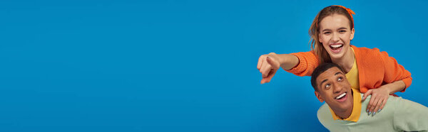 banner, joyful african american man piggybacking girlfriend on blue background, pointing at camera