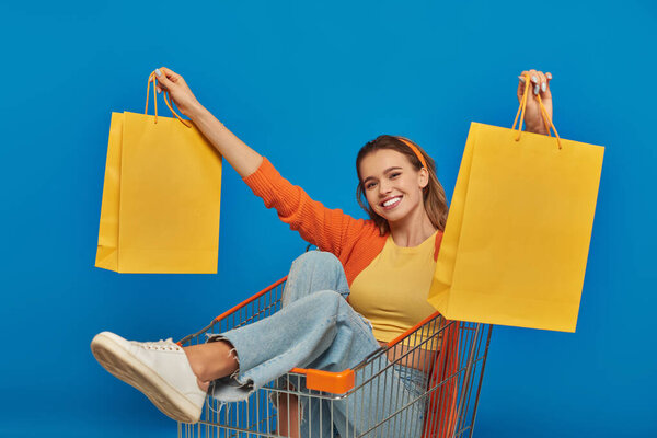 positive young woman sitting in cart and holding shopping bags on blue background, buying spree