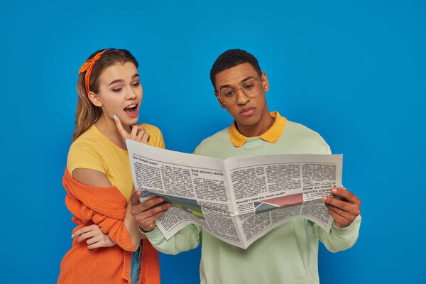 amazed woman reading newspaper near calm african american man in eyeglasses on blue backdrop