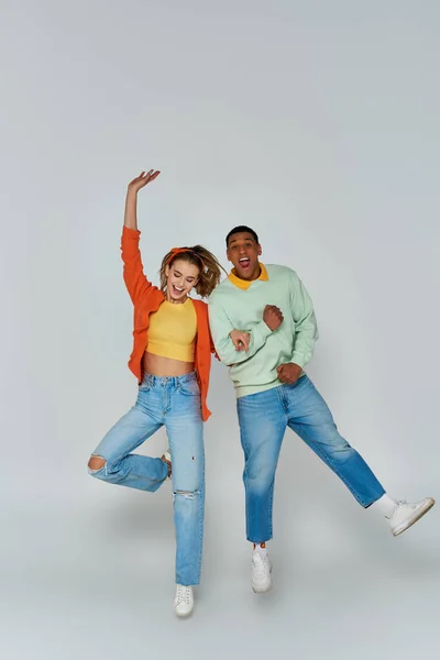 cheerful interracial couple in casual attire jumping and looking at camera on grey backdrop, fun