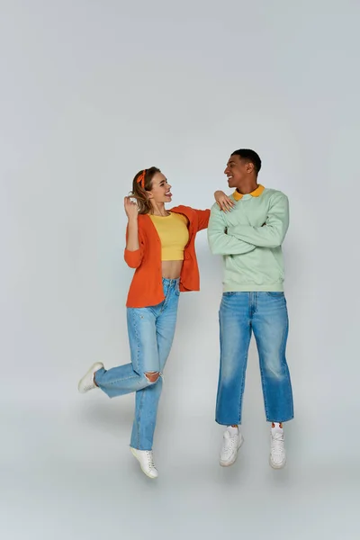 stock image happy young woman sticking out tongue and looking at african american man on grey backdrop, levitate