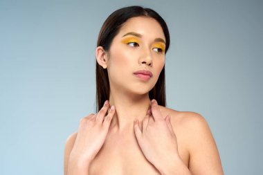 pretty model with bold makeup and bare shoulders posing on blue backdrop, asian beauty concept clipart
