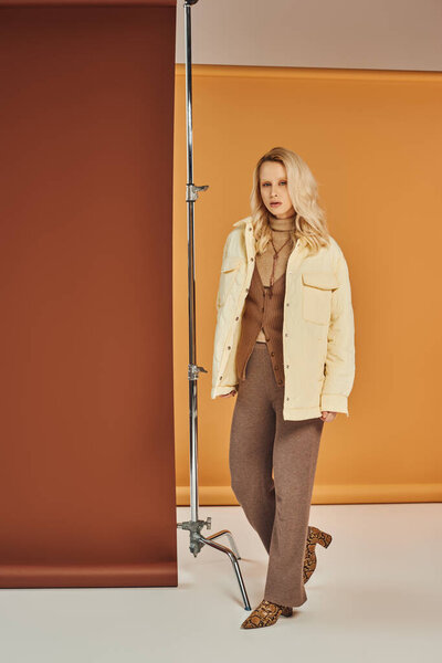 stylish woman in outerwear and boots looking at camera while walking on pastel backdrop, fashion