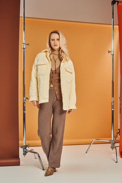 блондинка в стильной верхней одежде и сапогах, смотрящая в камеру и позируя в студии, осенняя мода