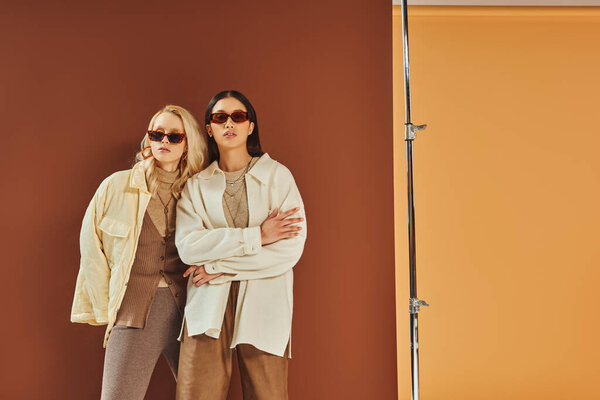 Осенняя мода и тенденции, межрасовые женщины в солнцезащитных очках и верхней одежде позируют на фоне цвета дуэта