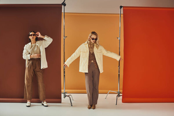 азиатка в солнцезащитных очках и осенней верхней одежде позирует рядом с моделью на красочном фоне, осенней моде