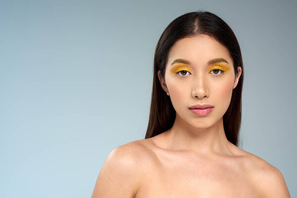 asian woman with brunette hair and bold makeup looking at camera isolated on blue, diverse beauty