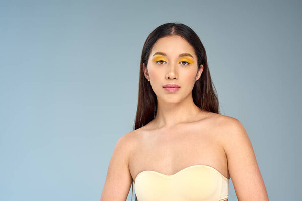 asian model with bold makeup posing in strapless top isolated on blue, diverse beauty and eye makeup