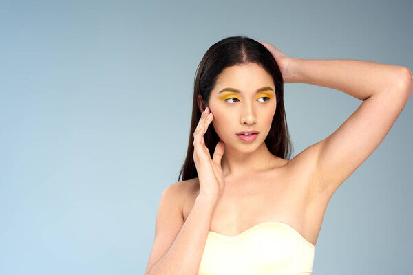 asian woman with bold makeup posing in strapless bra on blue backdrop, radiant skin and visage