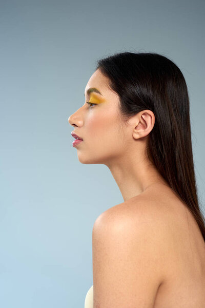 side view of model with bold makeup and bare shoulders posing on blue backdrop, asian beauty concept