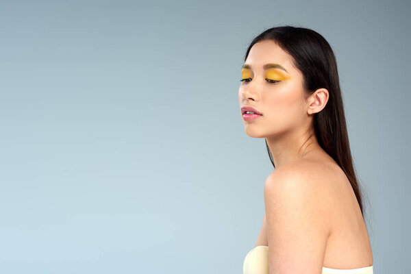 beauty, asian model with bold makeup and bare shoulders posing on blue backdrop, female elegance