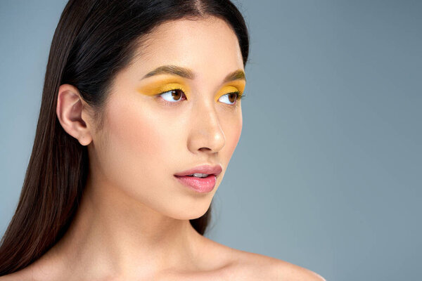 гламурная азиатская женщина с смелым макияжем и обнаженными плечами, отворачивающаяся и позирующая на синем фоне