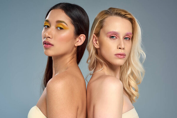 interracial models with vibrant eye makeup looking at camera on blue backdrop, beauty trend concept