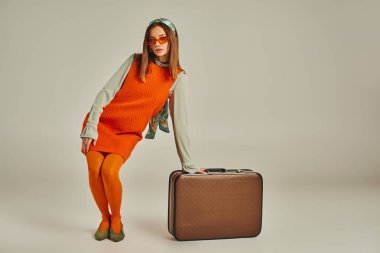 Turuncu elbiseli çekici bir kadın. Gri bavulun yanında poz veriyor. Geçmişten kalma moda.