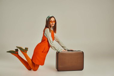Eski moda kıyafetler ve güneş gözlüklü genç bir kadın gri bavulun yanında diz çöküyor.