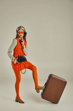 young old-fashioned woman in sunglasses talking on retro rotary phone near vintage suitcase on grey clipart