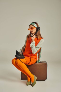 trendy retro-style woman sitting on vintage suitcase and talking on corded phone on grey backdrop clipart