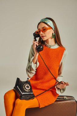Güneş gözlüklü retro-ilham verici kadın, gri bavulun üzerinde otururken telefonla konuşuyor.