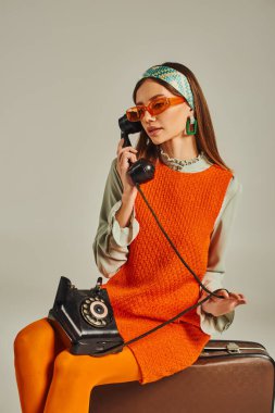 retro-inspired woman in sunglasses talking on dial phone while sitting on vintage suitcase on grey clipart