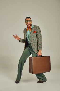Ekose ceketli, güneş gözlüklü, gri, eski moda bir valizle poz veren neşeli bir adam.