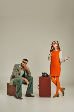 Turuncu elbiseli kadın telefonla konuşuyor. Sıkılmış bir adamın yanında. Gri bavulun üzerinde oturuyor.