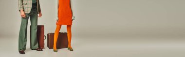 Gri pankartın yanında turuncu elbiseli ve ekose ceketli eski moda bavullu çift manzarası.