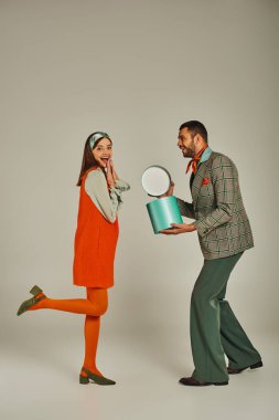 Tarz sahibi bir adam, aşırı neşeli turuncu elbiseli kadının yanında hediye kutusu tutuyor. Gri, retro-esinlenmiş moda.
