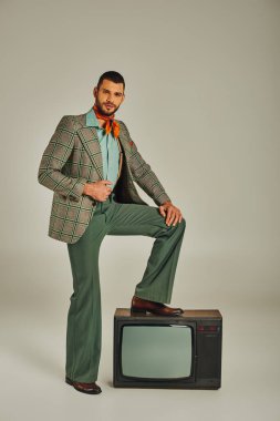 Kendine güvenen, modaya uygun retro elbiseli, klasik televizyona basan ve gri kameraya bakan bir adam.