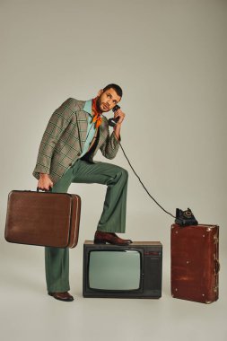 Modayı takip eden bir adam bavulu tutuyor ve eski moda TV setinin yanındaki eski moda telefon kulübesinde konuşuyor.