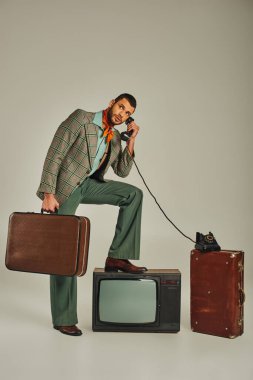 trendy man holding suitcase and talking on corded phone near vintage tv set on grey, retro lifestyle clipart