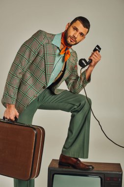 Elinde bavulu olan bir adam, gri, eski moda bir televizyonun yanında telefon ahizesi tutuyor.
