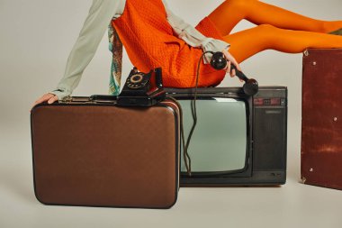 Turuncu elbiseli bir kadının eski bir bavula oturması ve gri, eski moda bir televizyona oturması.
