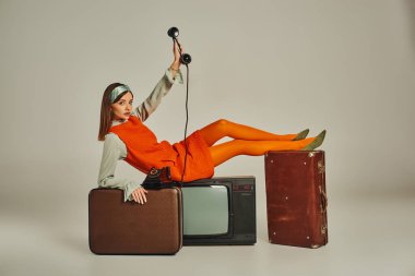 Modaya uygun retro giyimli genç bir kadın. Televizyonda antika bir telefon ve gri bavullarla oturuyor.
