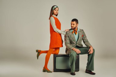 Turuncu elbiseli kadın, erkeğin omuzlarına dokunuyor. Gri, eski moda TV setinde oturuyor.