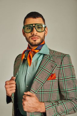 retro style man in plaid blazer and colorful neckerchief with sunglasses looking at camera on grey clipart