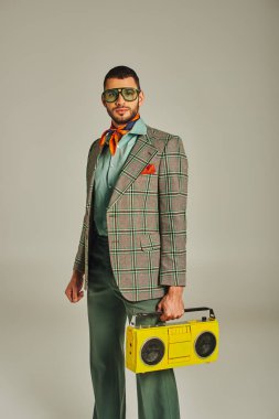 trendy man in plaid blazer and sunglasses holding yellow boombox on grey, retro-inspired lifestyle clipart