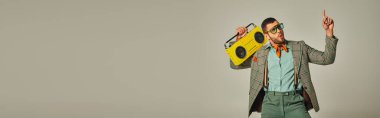 excited stylish man in sunglasses holding vintage boombox, pointing up and dancing on grey, banner clipart