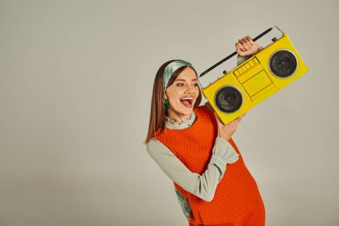 Turuncu retro tarzı elbisesi olan aşırı neşeli bir kadın sarı boombox taşıyor ve gri üzerinde dans ediyor. Mutluluk.