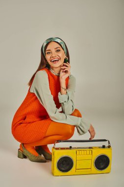 Parlak turuncu elbiseli mutlu bir kadın, gri, klasik titreşimli sarı müzik kutusunun yanındaki kameraya bakıyor.