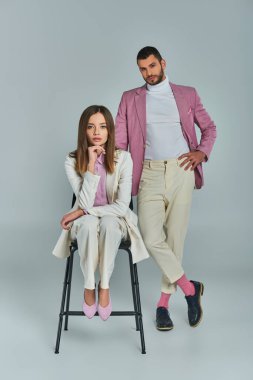 confident man in lilac blazer with hand on hip near woman in white suit sitting on chair on grey clipart
