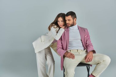 Beyaz takım elbiseli genç bir kadın, gri arka planda oturan leylak rengi ceketli kendine güvenen bir adama yaslanıyor.