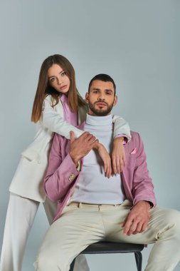 Beyaz takım elbiseli modacı kadın, gri arka planda oturan leylak ceketli, kendine güvenen adamı kucaklıyor.