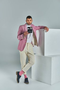 stylish man in lilac blazer and white pants taking photo on vintage camera near cubes on grey clipart