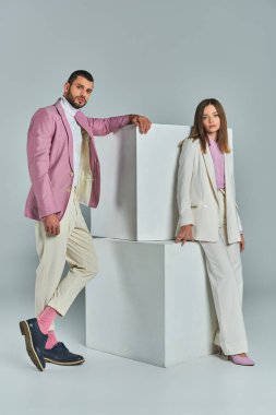 Gri, minimalist moda üzerine beyaz küplerin yanında poz veren şık takım elbiseli genç ve kendinden emin bir çift.